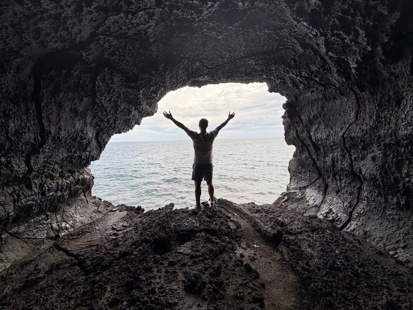 Figure framed by a lava sea cave with arms raised toward the ocean, awakening in the elements