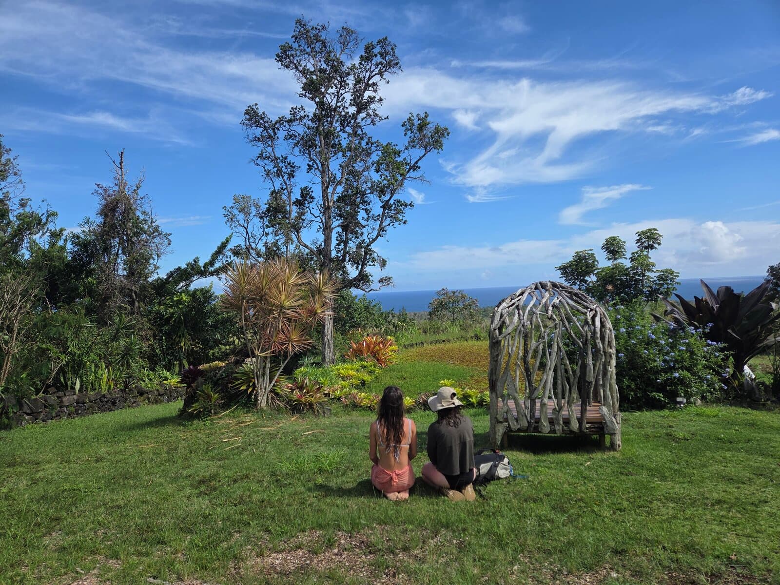 Two retreat participants sitting on the sanctuary lawn facing the ocean, a driftwood gazebo and tropical garden surrounding them